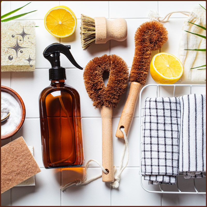 Flat lay of eco-friendly cleaning tools including wooden brushes, amber spray bottle, lemon slices, baking soda, and striped linen towels on white tile