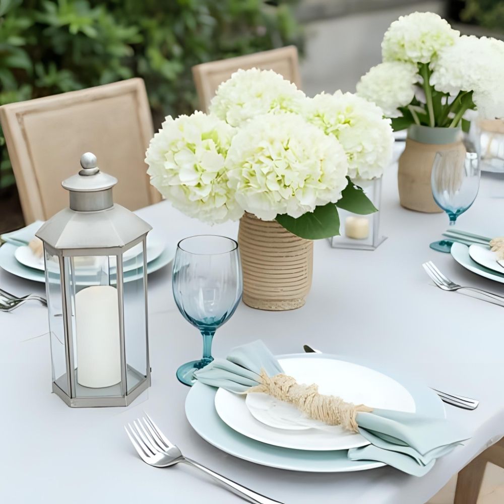 Coastal table setting with white hydrangeas, blue glassware, and hurricane lanterns