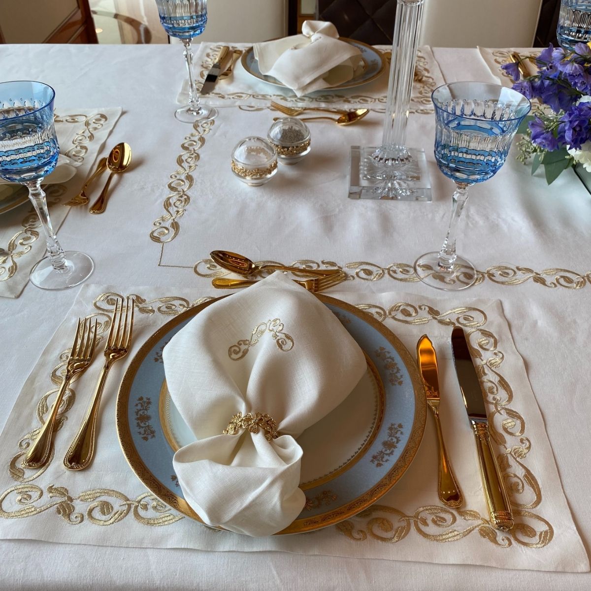 Luxury place setting with gold flatware, blue and gold rimmed plates, crystal glasses and embroidered linen napkin