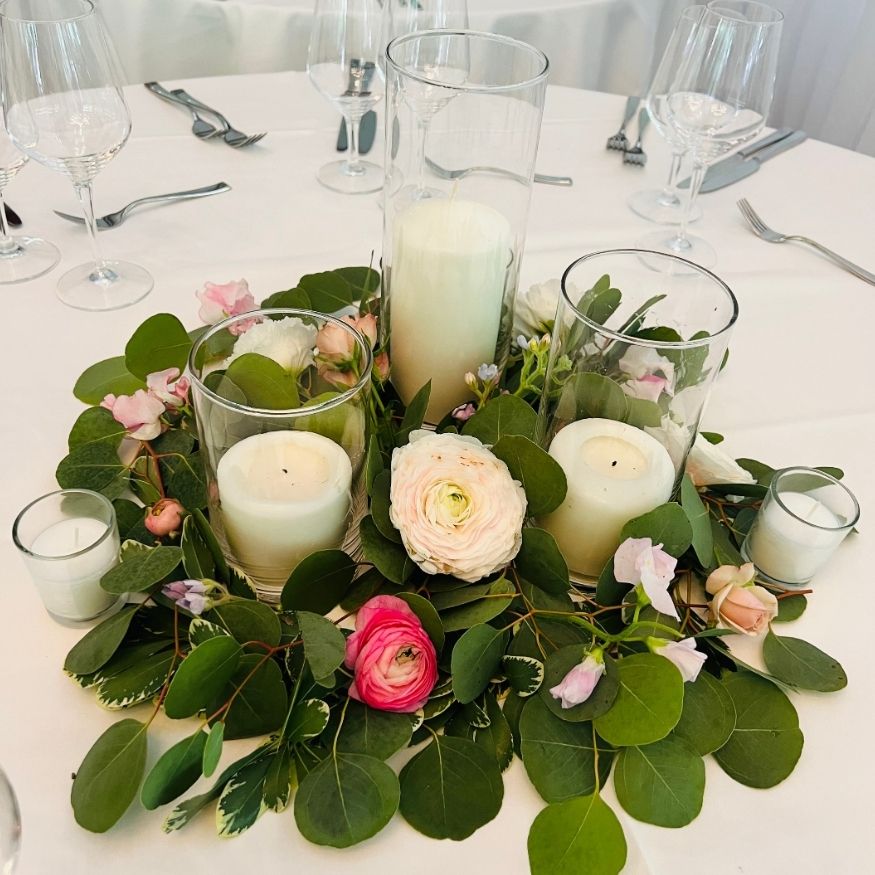 Low floral centerpiece with eucalyptus greenery, pink roses, and pillar candles in glass hurricane vases on dinner table