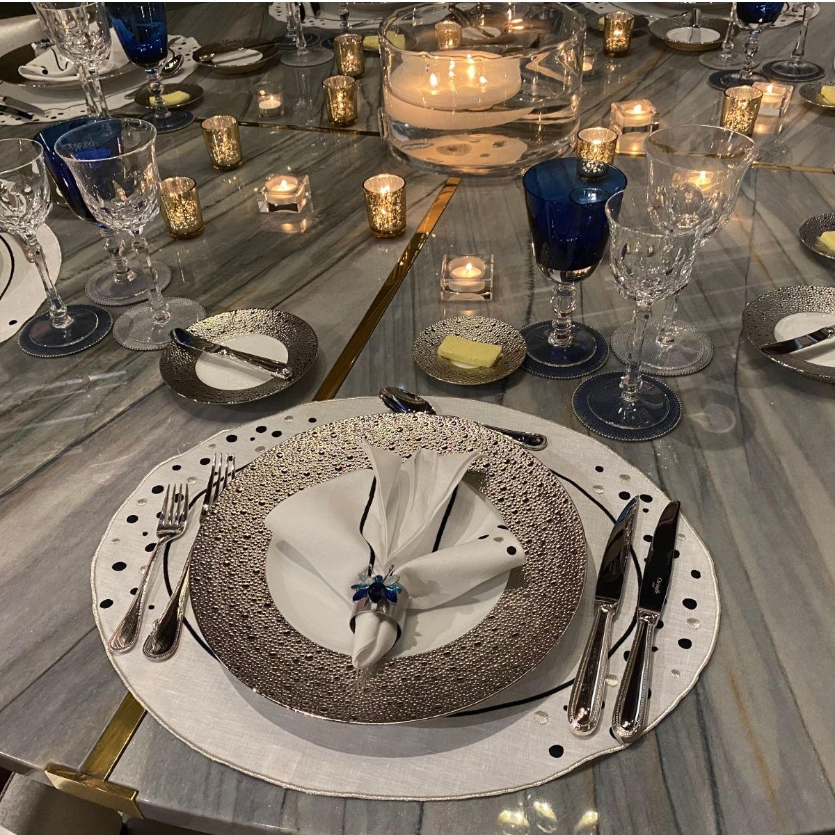 Modern glamorous table setting with marble table, silver chargers, blue goblets and candlelight