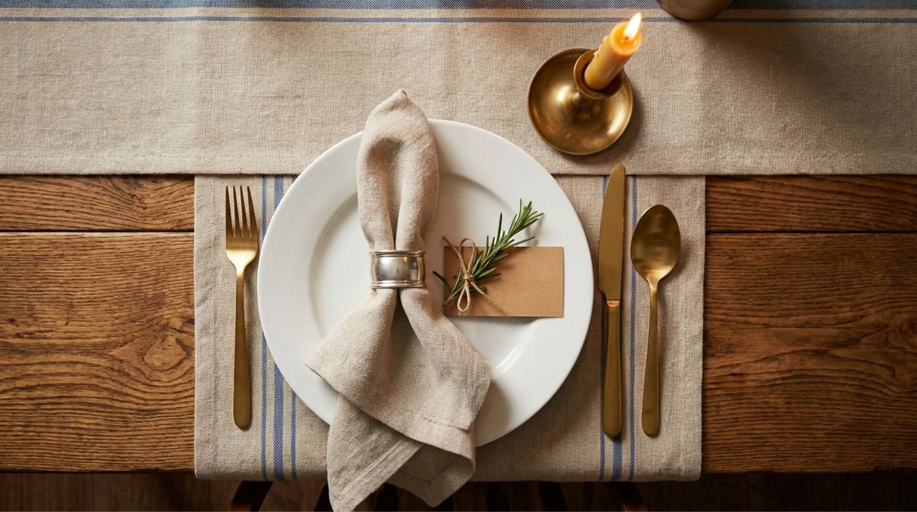 Everyday table setting with linen runner, cloth napkin with brass ring, rosemary sprig, and taper candle on wooden table
