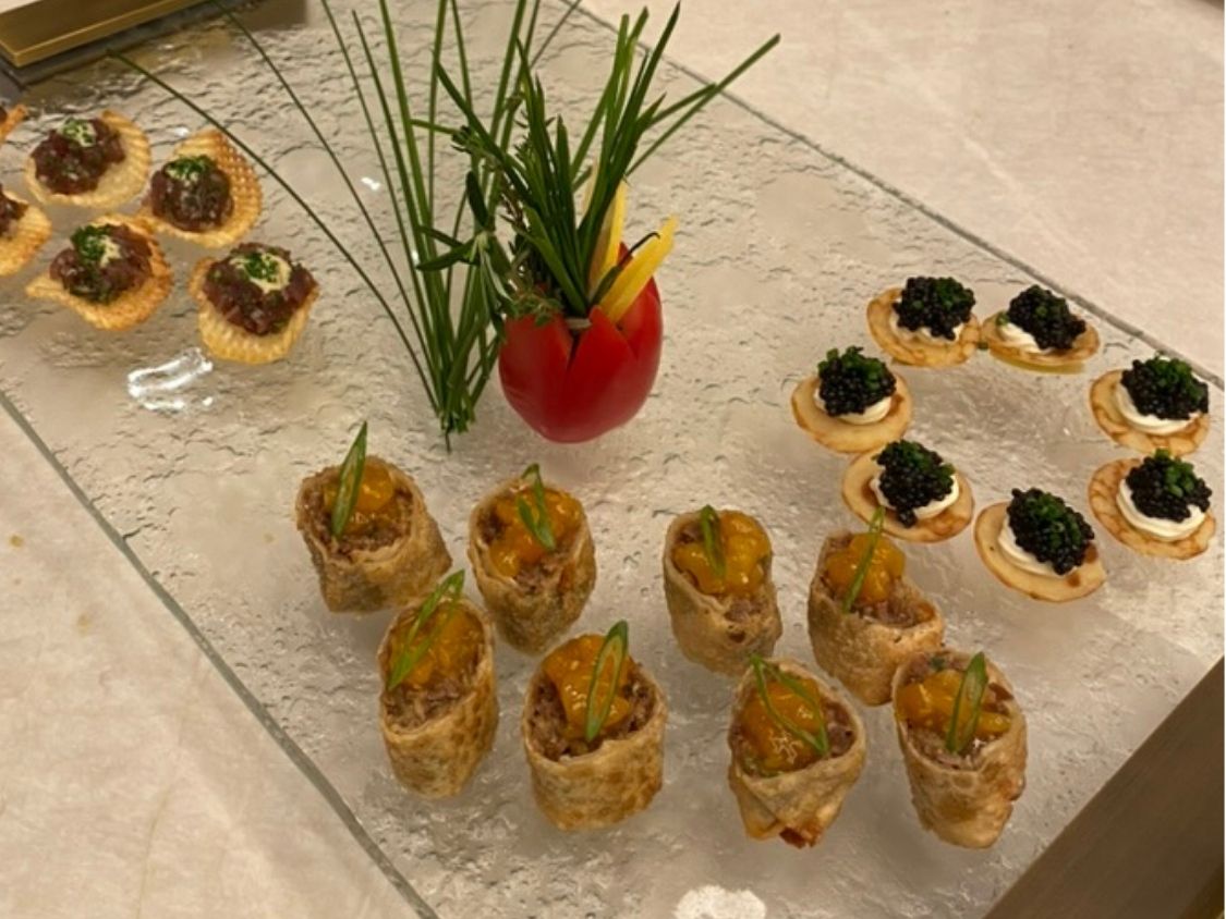 Glass serving tray with brass handles holding tartare crisps, caviar blini, and canapés with a carved tomato herb garnish