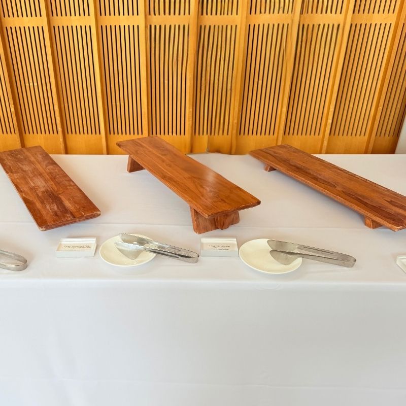 Three wooden serving boards and footed risers set on a white buffet table with serving tongs